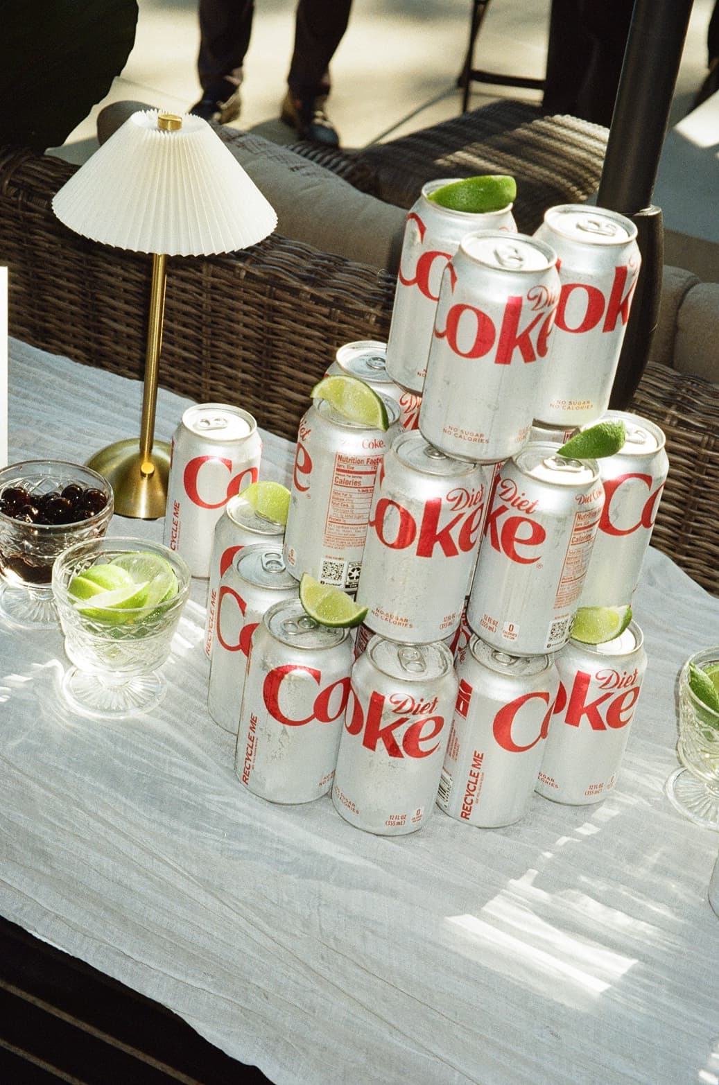 A pyramid of Diet Coke cans topped with lime wedges on a linen-covered patio table, beside a brass lamp and glass bowls of cherries and limes